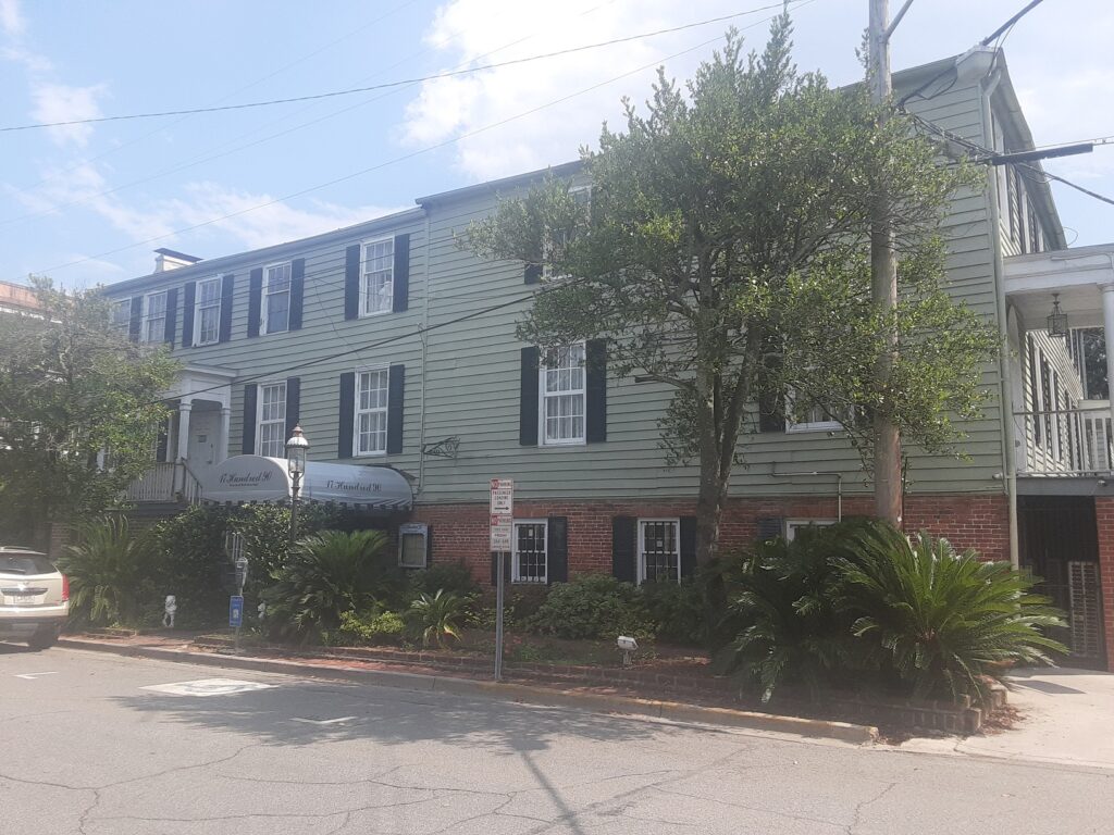 A long green clapboard inn with black shutters on a brick base, behind palms and a street tree