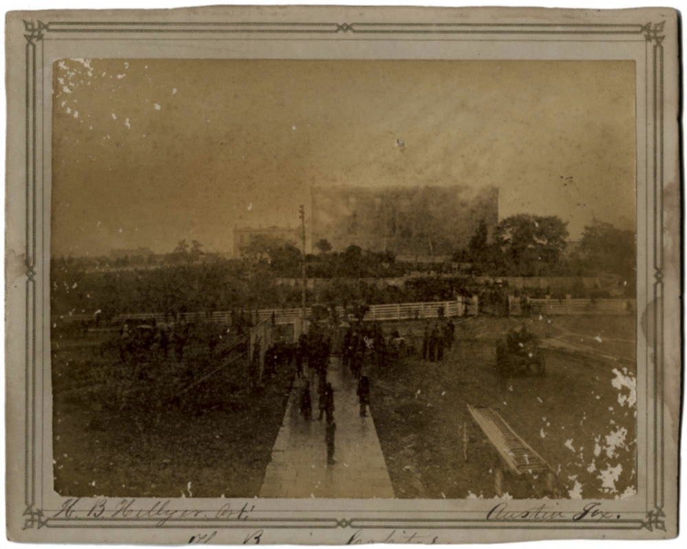 A faded 1881 photograph of a crowd on open ground watching smoke rise from the old Texas Capitol