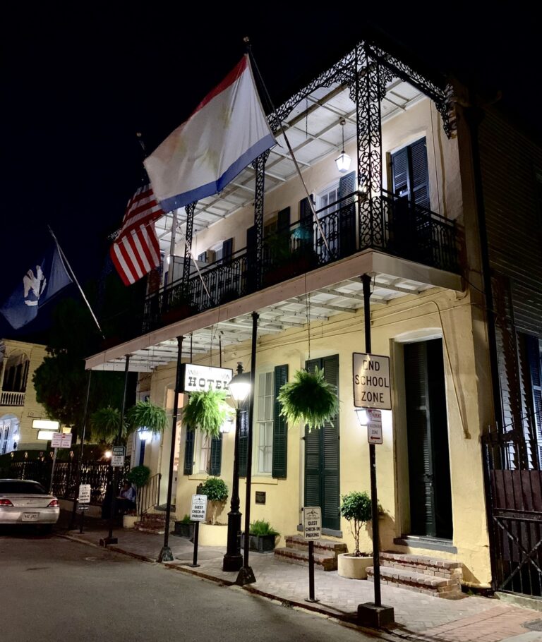 A lit two-story building with iron balconies, hanging ferns and flags, at night