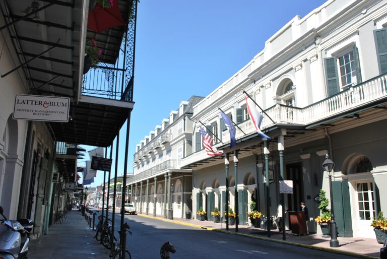A long white hotel front with green shutters, balconies and flags along a narrow French Quarter street