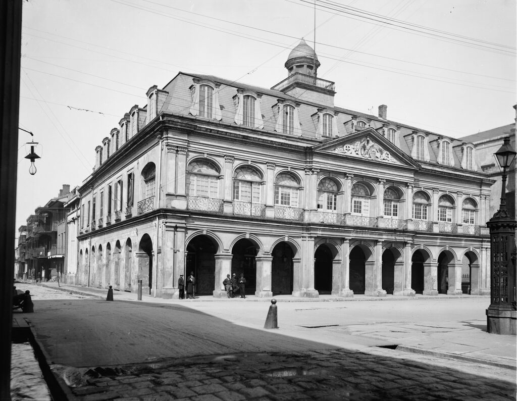 A two-story arcaded stone building with a mansard roof and a cupola beside an empty square, in black and white