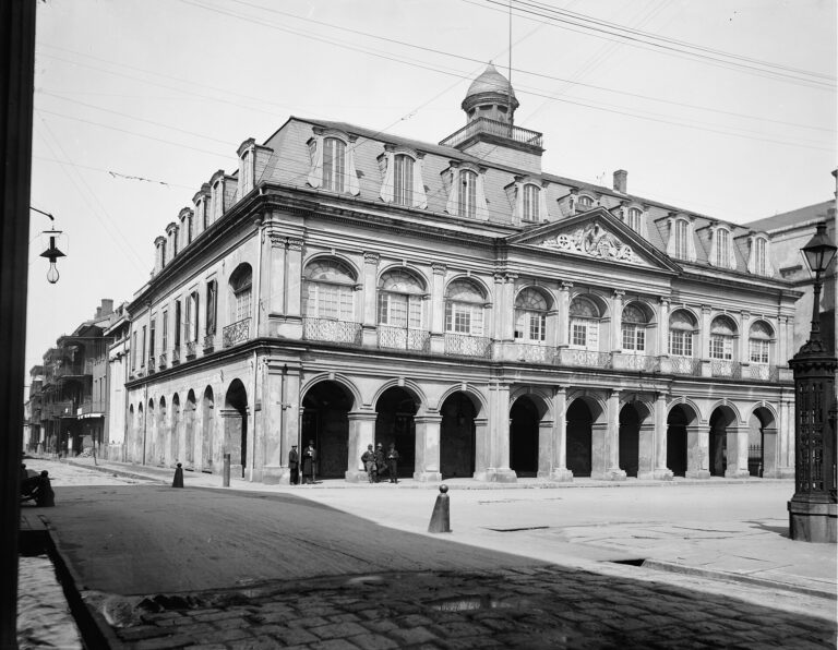A two-story arcaded stone building with a mansard roof and a cupola beside an empty square, in black and white