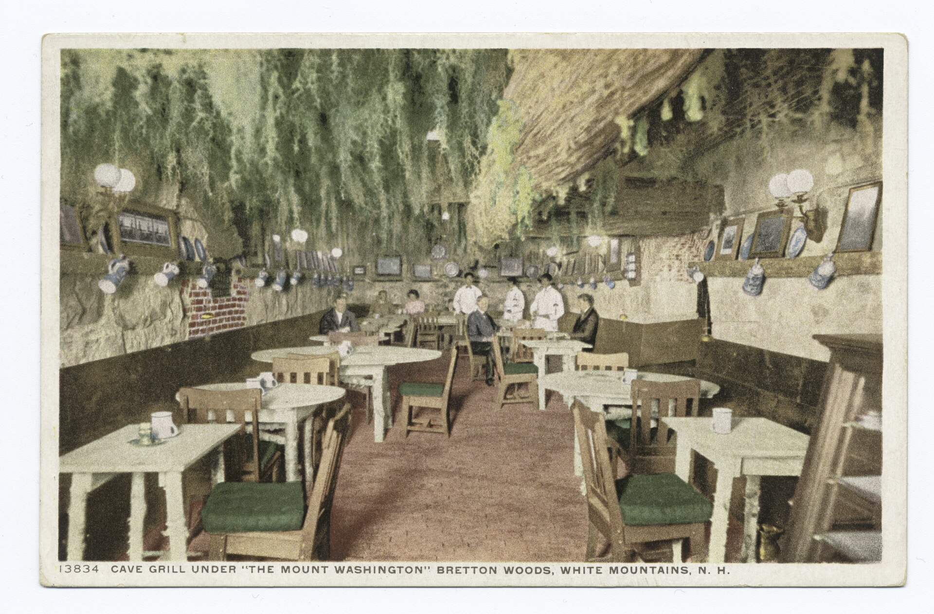 Tables and chairs in a low room with rough rock walls and hanging greenery, in a hand-colored postcard