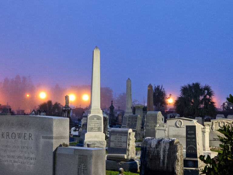 Tombs and an obelisk in a cemetery at dawn, with fog and streetlights behind