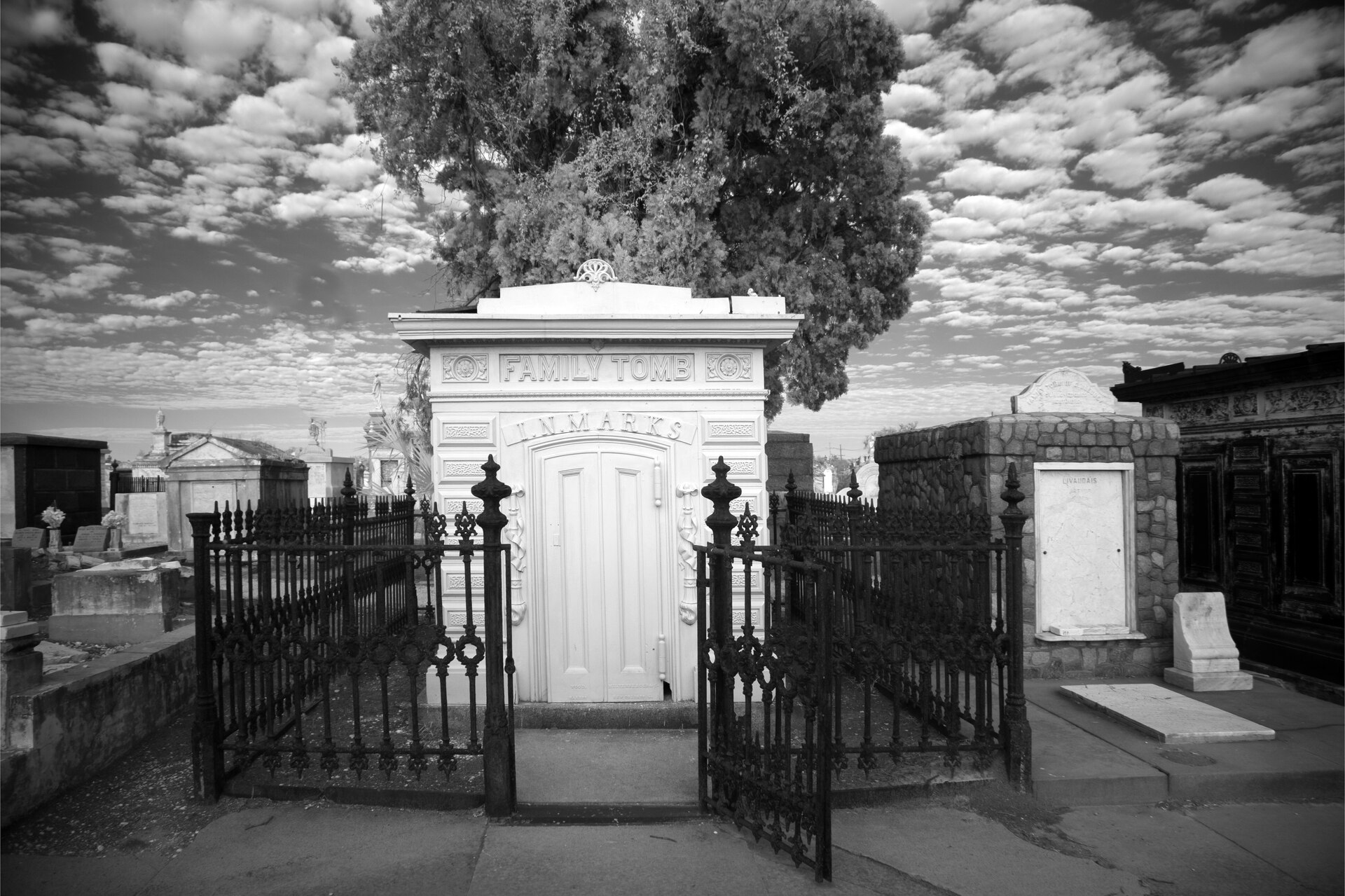 A black-and-white view of a white family tomb behind an open iron gate, under a clouded sky