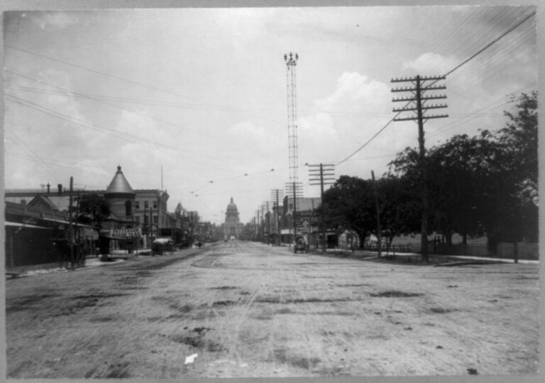 A wide, unpaved Congress Avenue about 1901, with a tall lattice tower in the middle of the street and the Texas Capitol dome in the distance