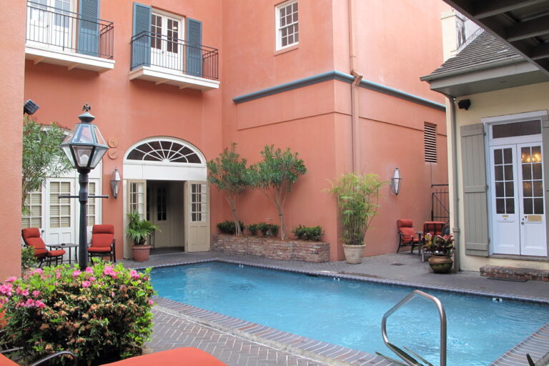 A small pool in a courtyard walled in salmon-pink stucco, with an arched doorway, a lamppost and potted trees