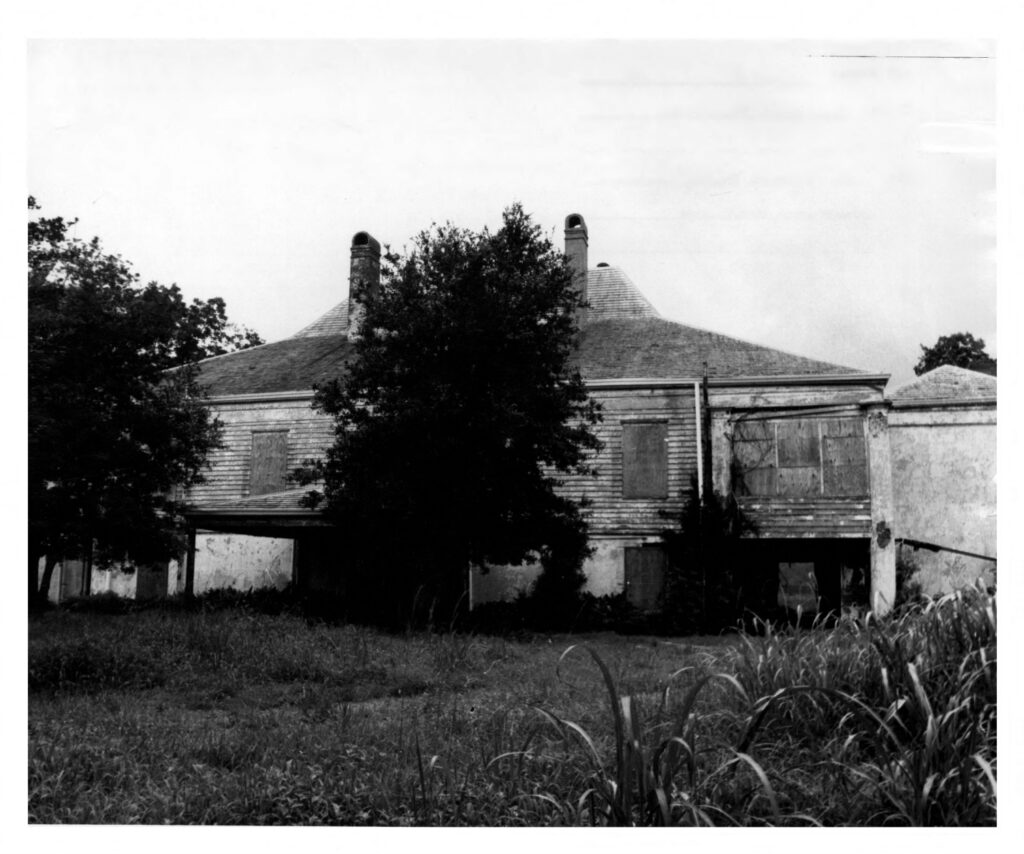 A broad two-story plantation house with a hipped roof and twin chimneys behind trees and tall grass, in black and white