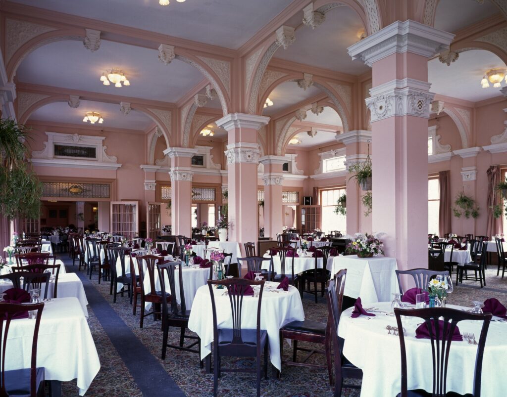 A long dining room with arched columns, chandeliers and tables set with white linen