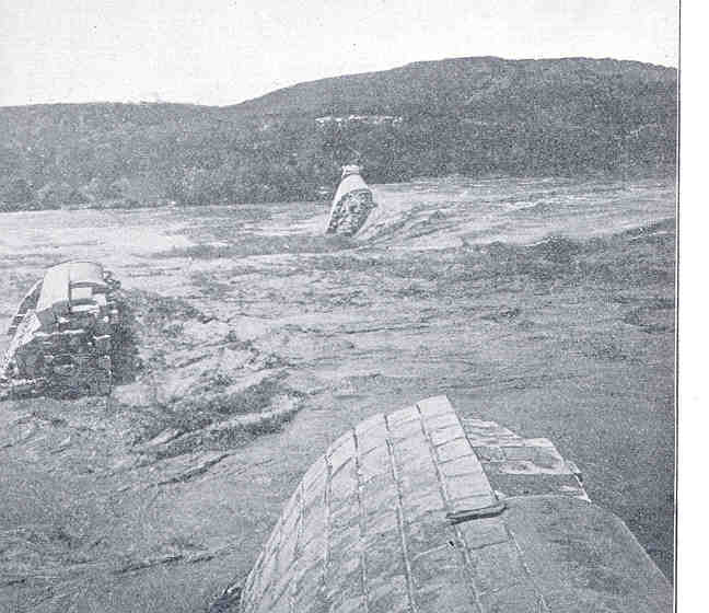 Black-and-white view of floodwater pouring past broken blocks of the Austin Dam, with hills behind
