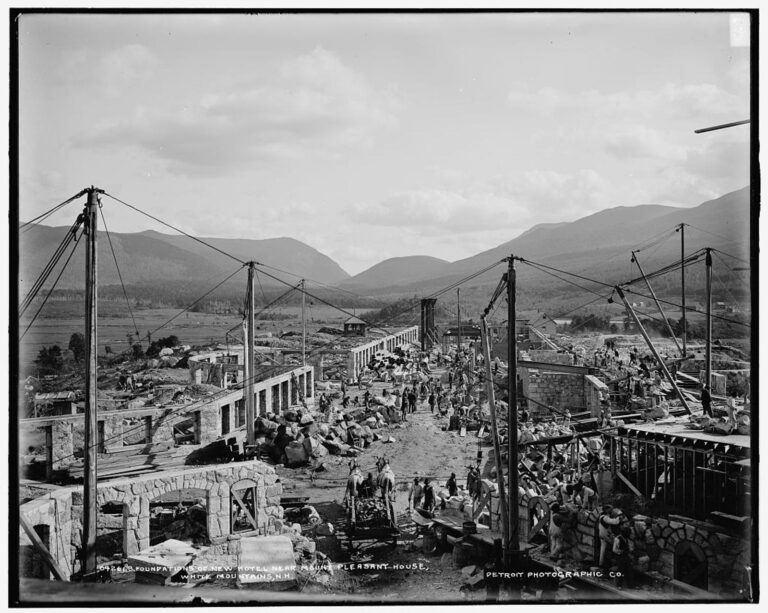 Stone foundation walls and wooden derricks across a construction site, with mountains behind