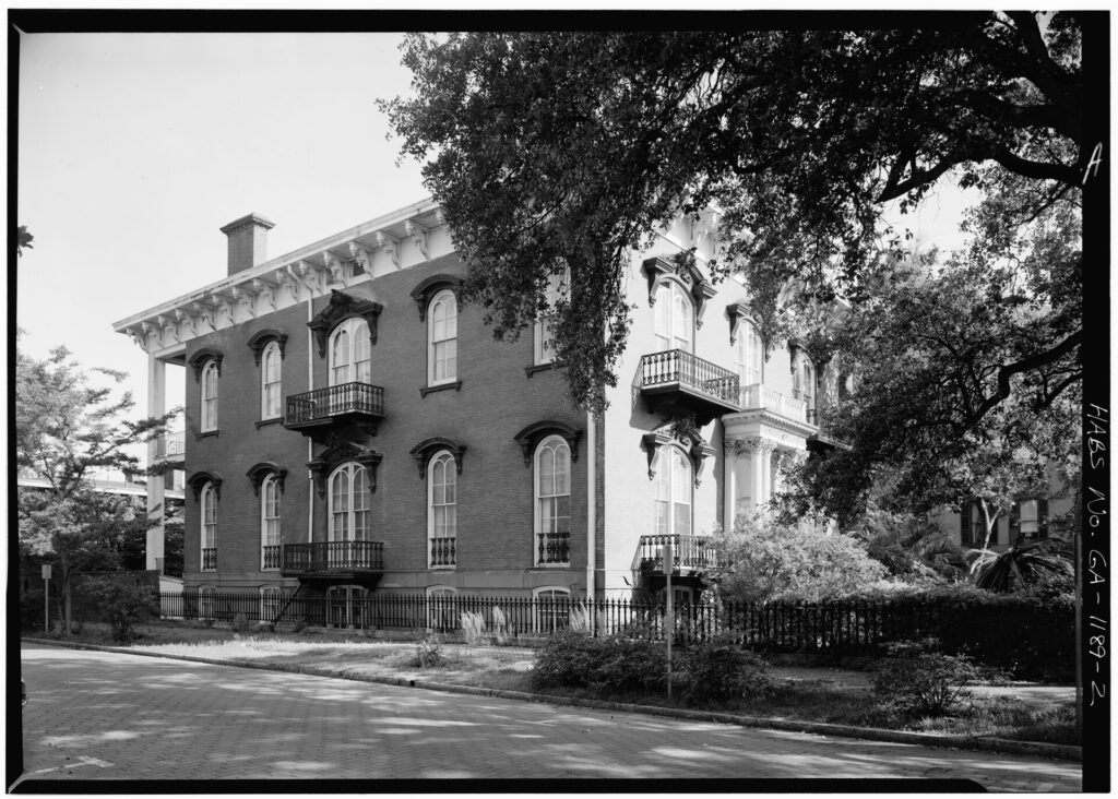 A black-and-white photo of a two-story brick mansion with arched windows and iron balconies behind live oaks