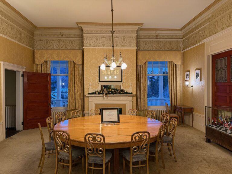 A round wooden table ringed by chairs under a chandelier, in a gold-papered room with a fireplace