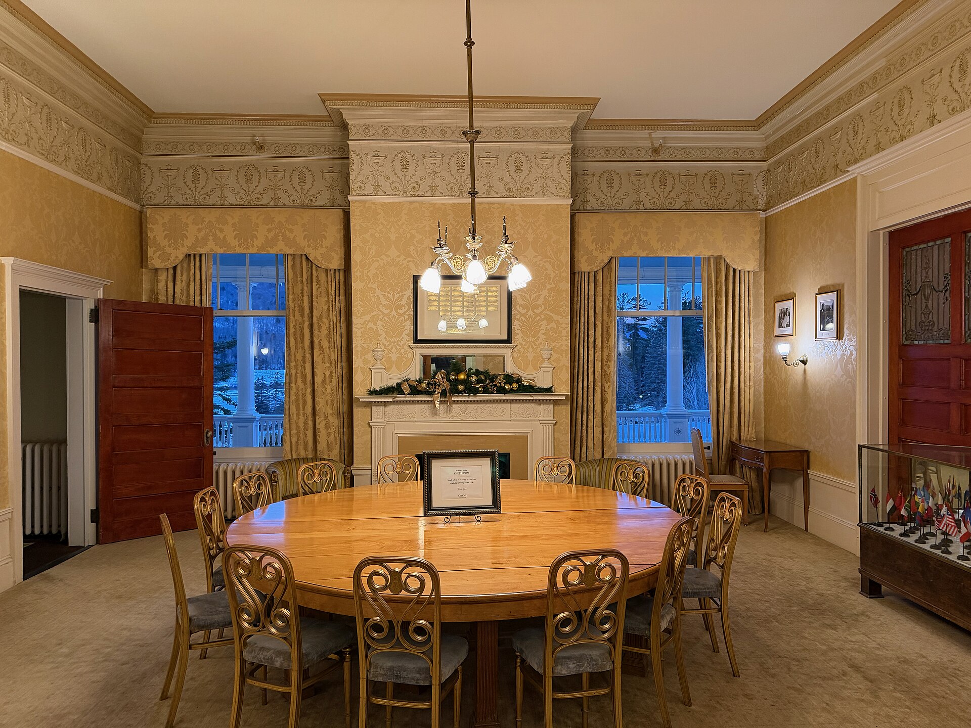 A round wooden table ringed by chairs under a chandelier, in a gold-papered room with a fireplace