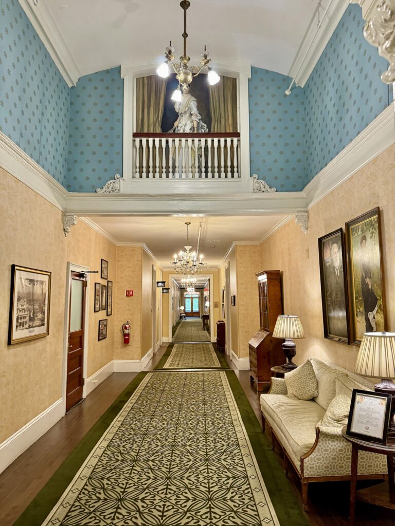 A long hallway with a patterned runner, armchairs and framed pictures, under a chandelier and a balcony railing