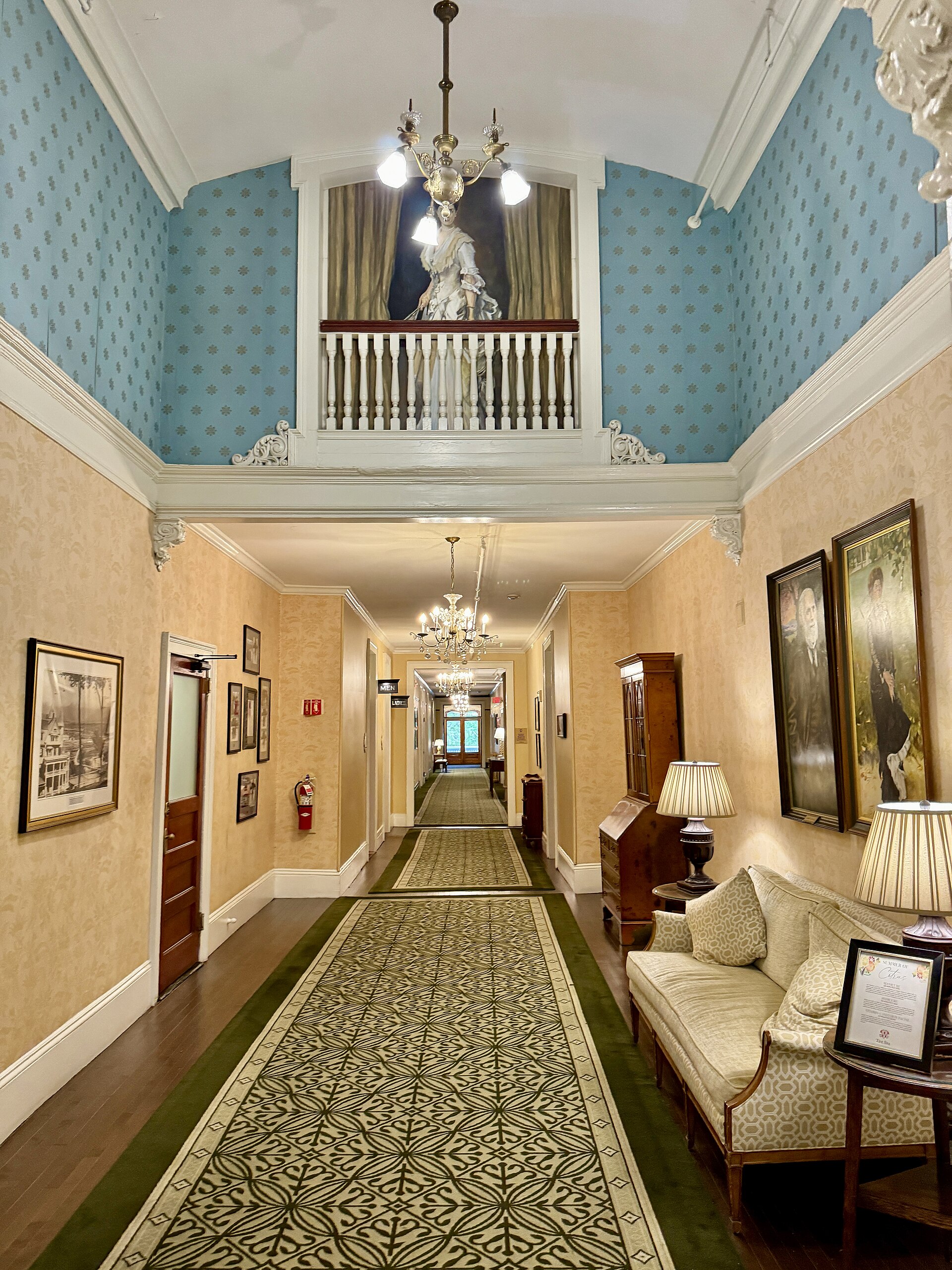 A long hallway with a patterned runner, armchairs and framed pictures, under a chandelier and a balcony railing