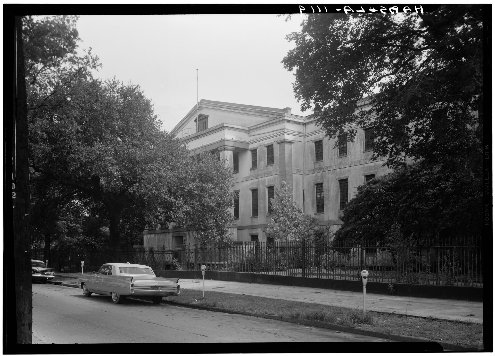 A large columned building behind trees and an iron fence, with a 1960s car parked in front
