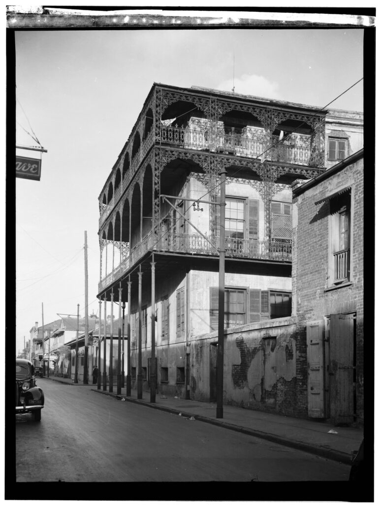 A tall stuccoed corner house wrapped in two levels of lacy cast-iron galleries, on an empty street