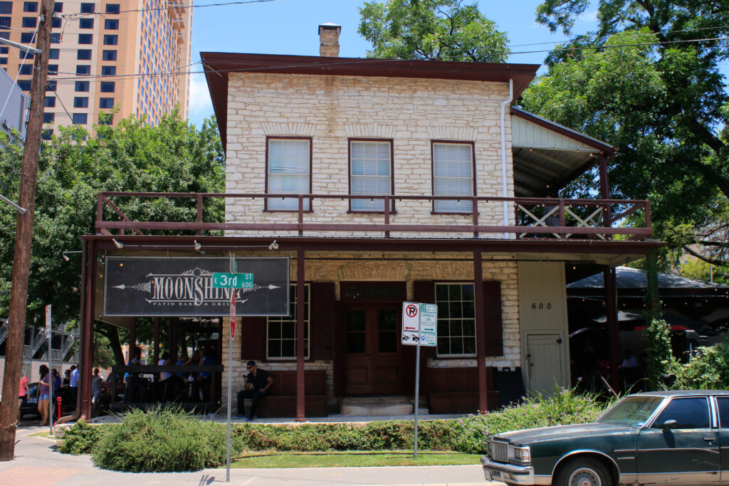 A two-story limestone store with a wooden porch and a Moonshine sign at a downtown Austin street corner