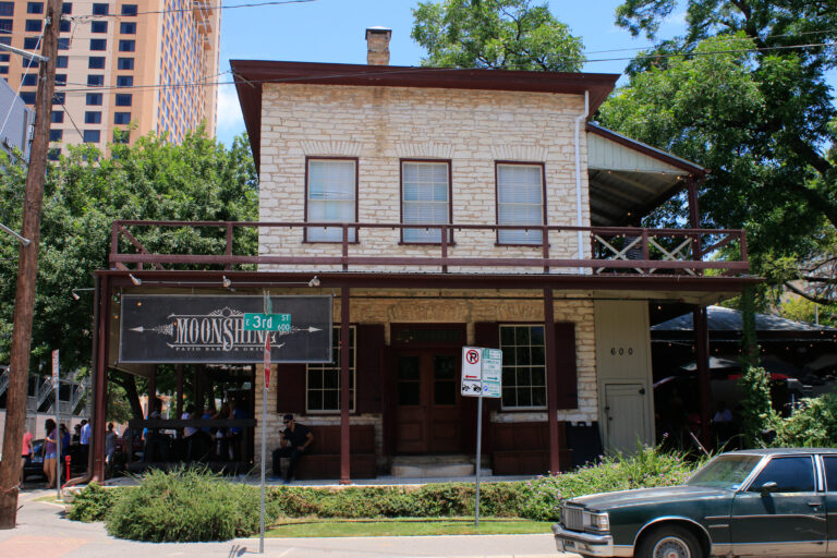 A two-story limestone store with a wooden porch and a Moonshine sign at a downtown Austin street corner