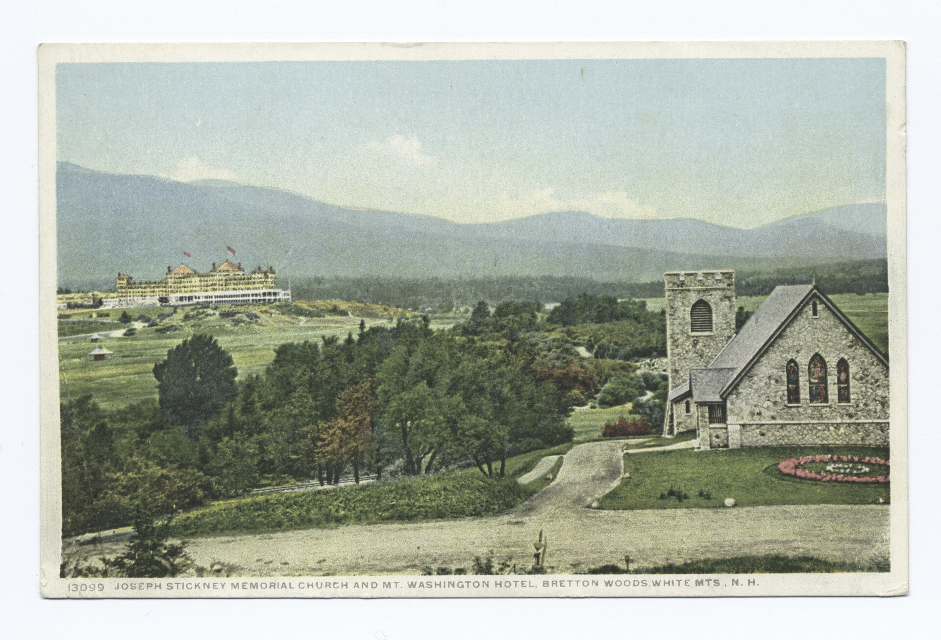 A small stone church with a square tower in the foreground, and the long hotel with red-flagged towers across a green valley