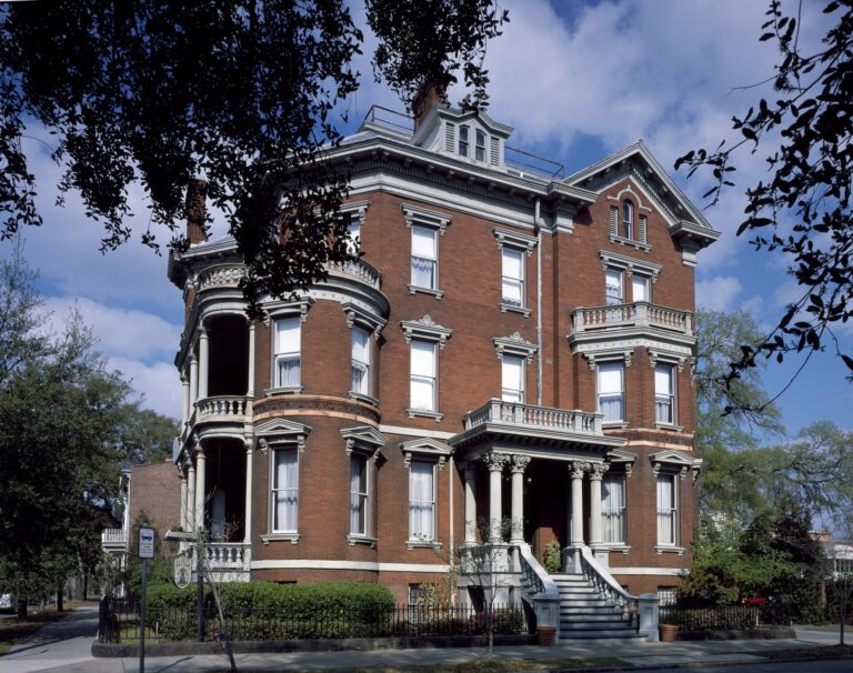 A red-brick Victorian mansion with white trim, curved porches and a tall front stair, framed by oak branches