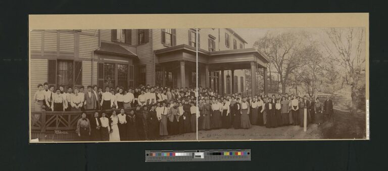 Rows of young women in long skirts and white blouses posed in front of a large wooden school building with a columned porch