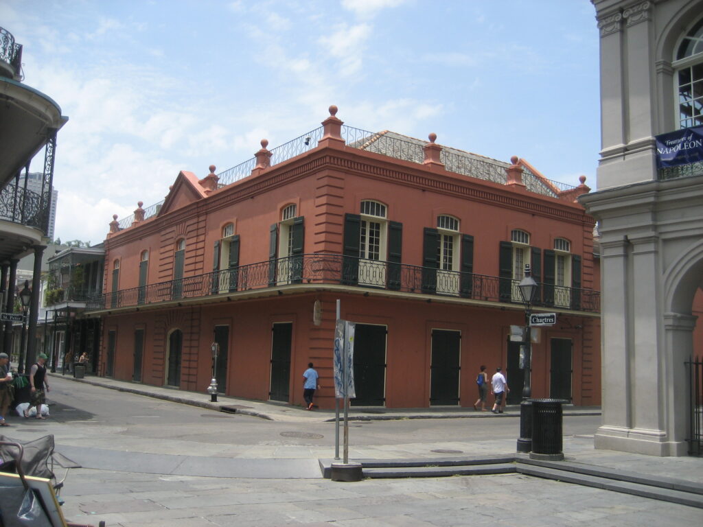 A salmon-pink two-story corner building with iron balconies and green shutters, beside the Cabildo's arches