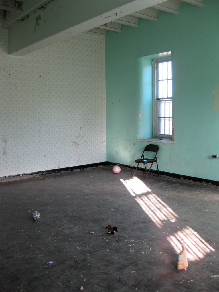 An empty room with mint-green and white walls, sunlight from a tall window falling across the floor, where a few balls and small toys lie near a folding chair