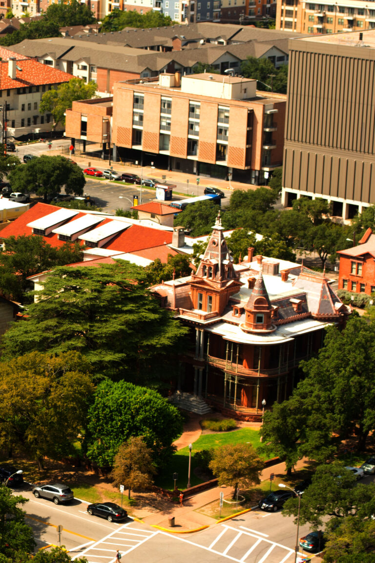 A turreted red Victorian house seen from above among trees and modern university buildings