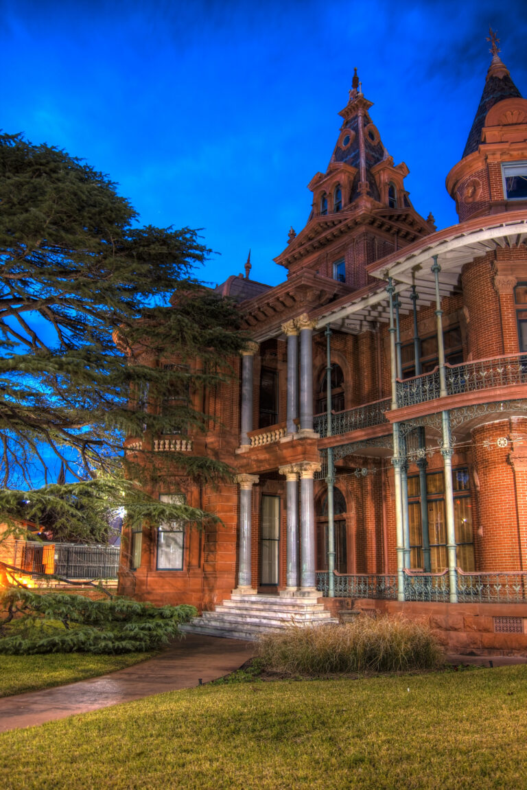 A red-brick Victorian mansion with turrets and two-story iron porches under a deep blue evening sky