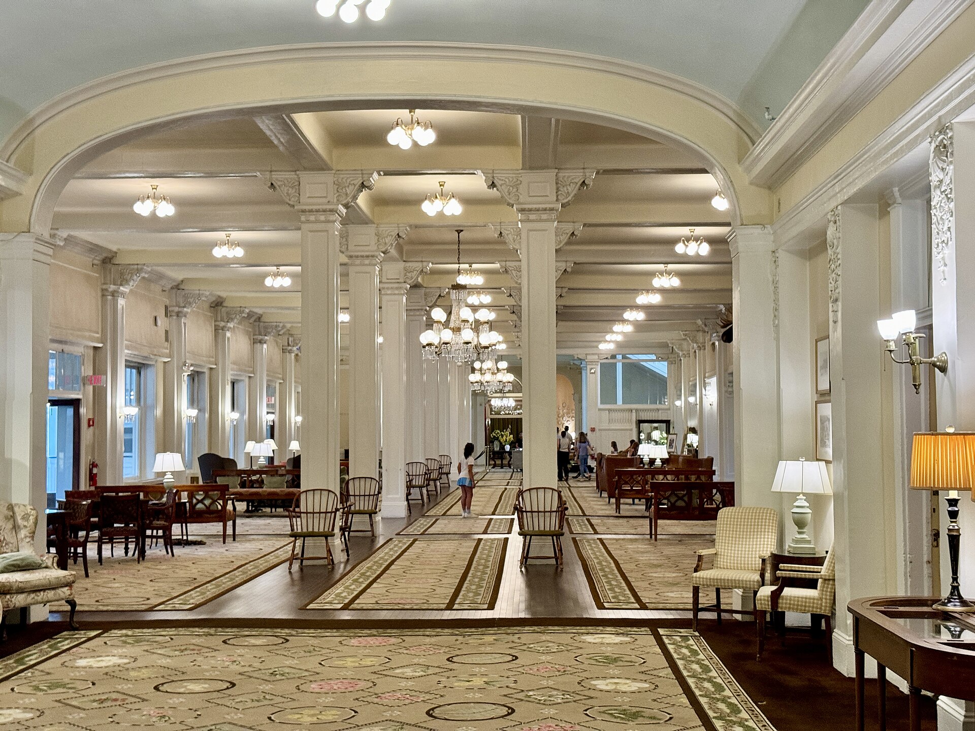 A long lobby lined with columns and chandeliers, with armchairs on patterned carpet