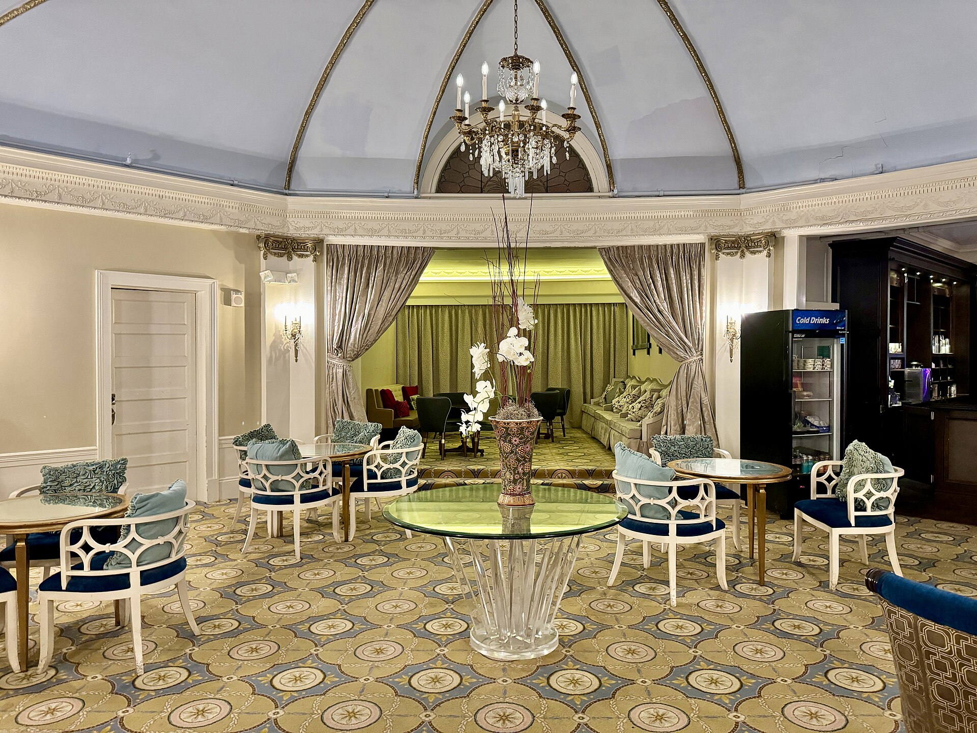 A round lounge under a ribbed, pale blue dome and a chandelier, with curtains drawn back from an alcove at the far end