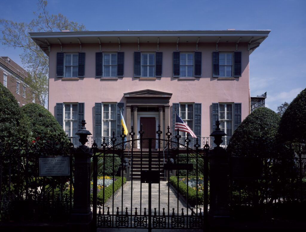 A pink stucco Italianate house with dark shutters behind an iron gate and clipped shrubs, flags by the door
