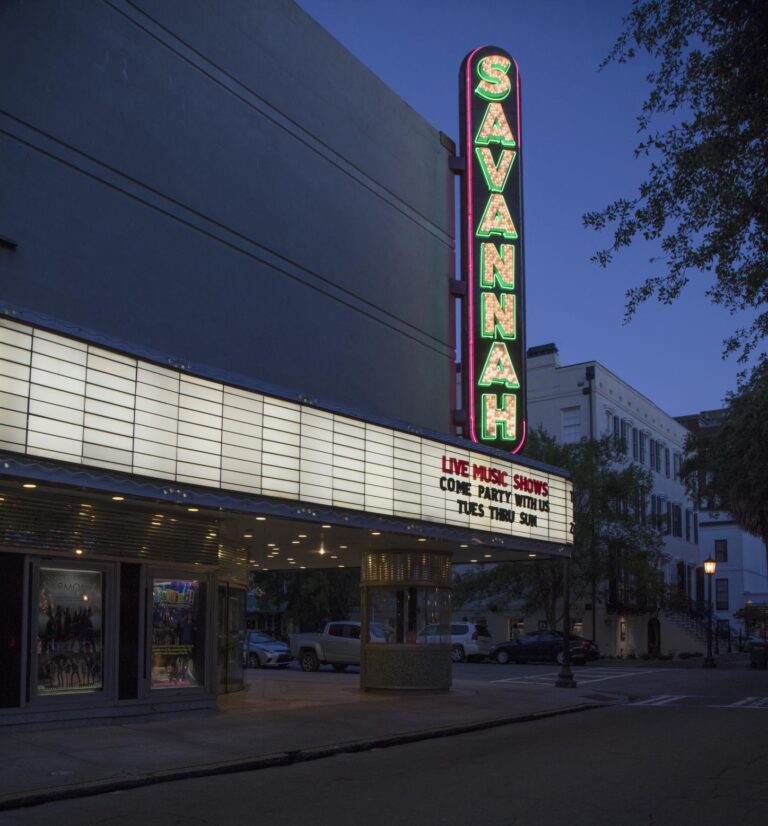 A vertical neon sign spelling SAVANNAH above a lit marquee on a plain theater front at dusk