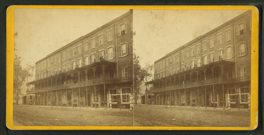 A sepia stereograph card with two images of a four-story hotel with long iron galleries over a dirt street