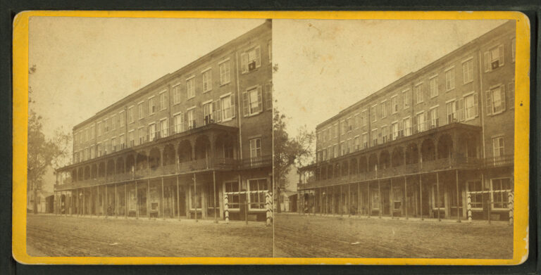 A sepia stereograph card with two images of a four-story hotel with long iron galleries over a dirt street