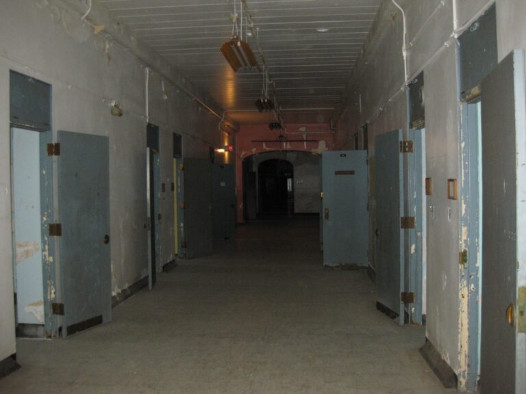 A long, dim hallway lined on both sides with open steel doors, paint peeling from the walls and ceiling