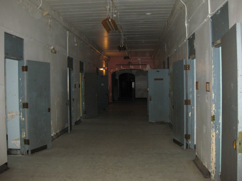 A long, dim hallway lined on both sides with open steel doors, paint peeling from the walls and ceiling