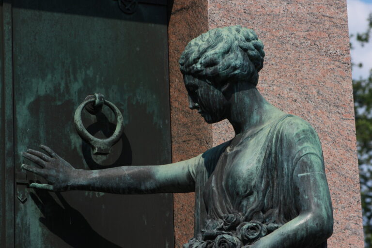 A green bronze statue of a woman reaching toward the ring knocker on a tomb door