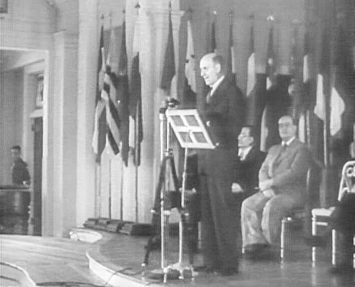 A man speaking at a lectern in front of a row of national flags, with officials seated beside him