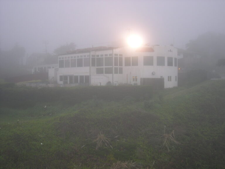 The white Moss Beach Distillery building on a grassy bluff in fog, with a light glowing above its roof