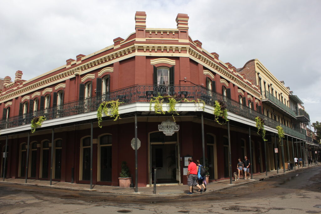 A three-story red-brick corner building with iron galleries hung with plants and a hanging sign reading Muriel's