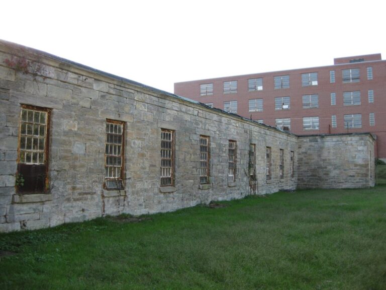 A long, one-story wing of rough stone blocks with tall barred windows beside a lawn, with a brick building behind it