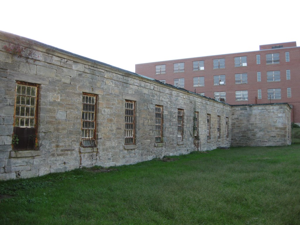 A long, one-story wing of rough stone blocks with tall barred windows beside a lawn, with a brick building behind it