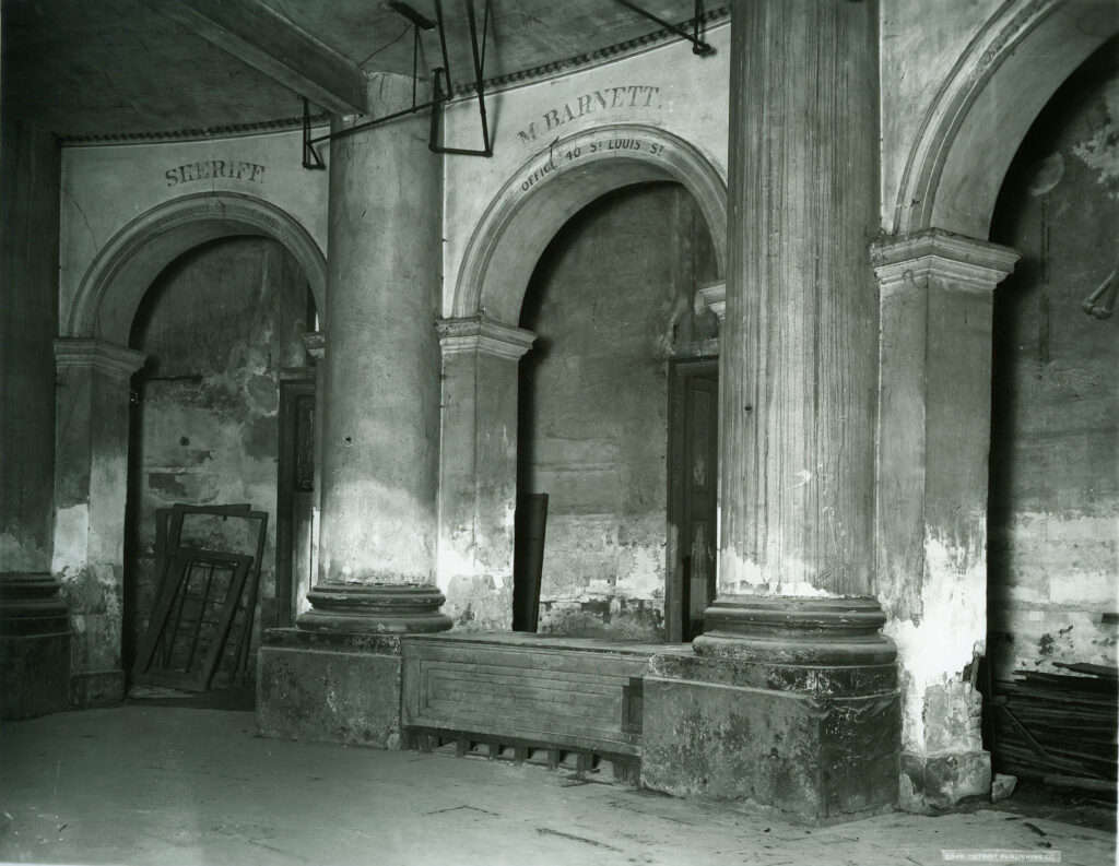 A dim black-and-white photo of a ruined arcade with columns, arches and a stone block between them