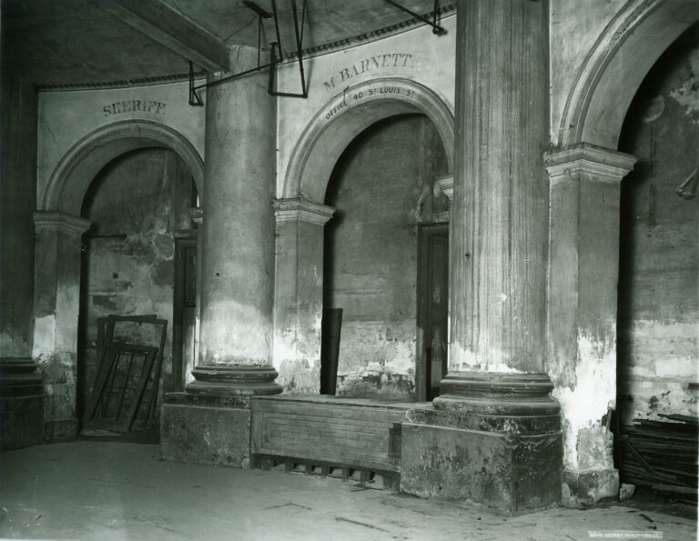 A dim black-and-white photo of a ruined arcade with columns, arches and a stone block between them