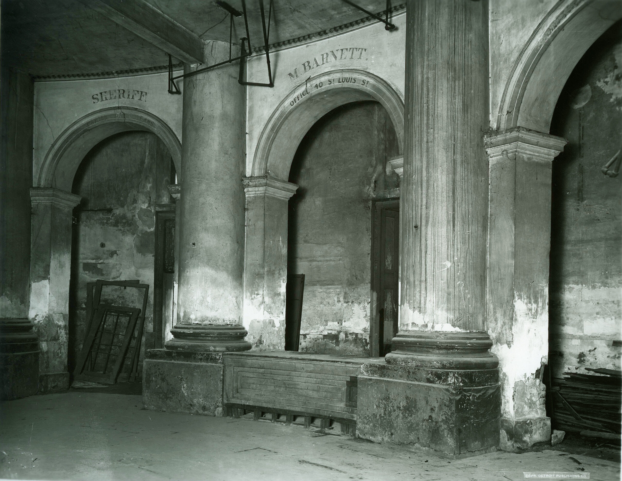 A dim black-and-white photo of a ruined arcade with columns, arches and a stone block between them