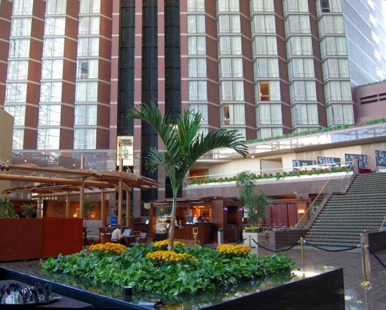 A glass-roofed atrium lobby with a planter of yellow flowers, a staircase, and floors of windows rising above