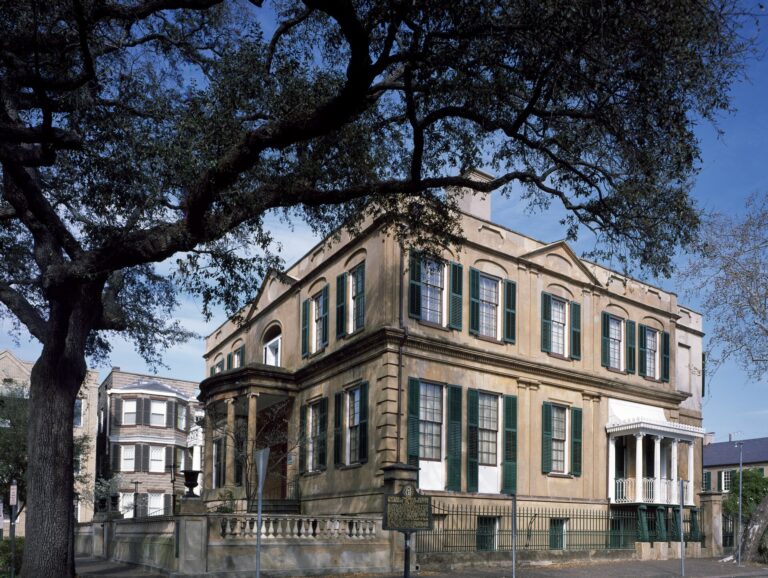 A cream stucco Regency mansion with green shutters, a curved porch and a balustrade under a spreading live oak